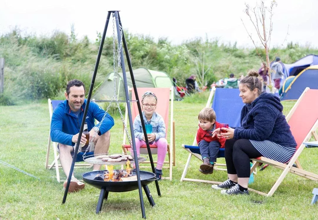 family using a bbq at a campsite with tents and people in the background